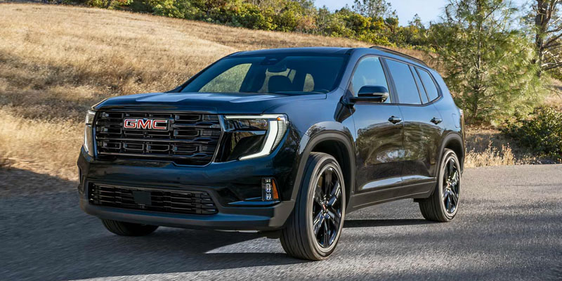 A dark black GMC SUV driving on a paved road next to a grassy hill.